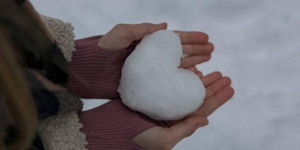 Close up of a woman's hands holding a heart sculpted in snow. You can see snow in the background as well.