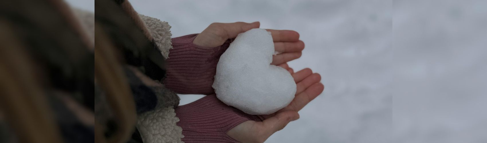 Close up of a woman's hands holding a heart sculpted in snow. You can see snow in the background as well.