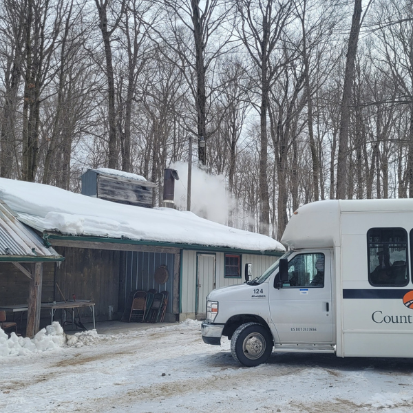 A maple syrup wooden building in the bush. It is winter, and there is a bus pulled up to the building. There is smoke coming out of the smokestack on the builiding, indicating they are boiling syrup inside.