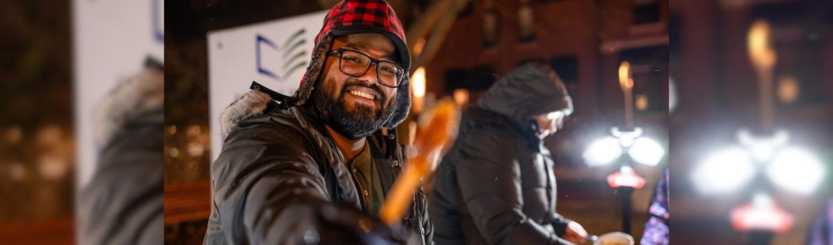 A smiling man wearing a plaid hat, winter jacket and smiling is reaching out to offer a stick with maple taffy on it. You can see another person working with maple syrup in the background.