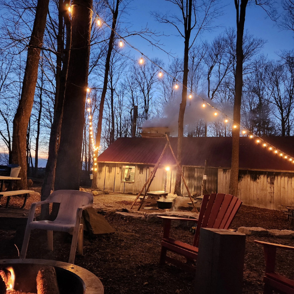 The maple bush shack at Sand Hills Maple Products in Wellesley. There is steam coming out of the stack in the building and lights strung around the outdoor area where you can see chairs, tables and a campfire area. It is twilight time in the winter. 
