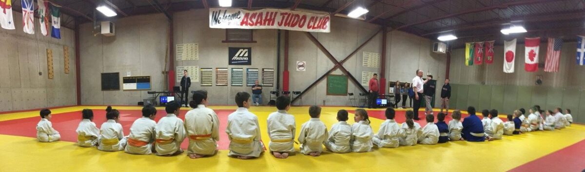 A group of students dressed in white judo uniforms seated on the floor of a gymnasium. There is a banner on the wall that read Asahi Judo Club.