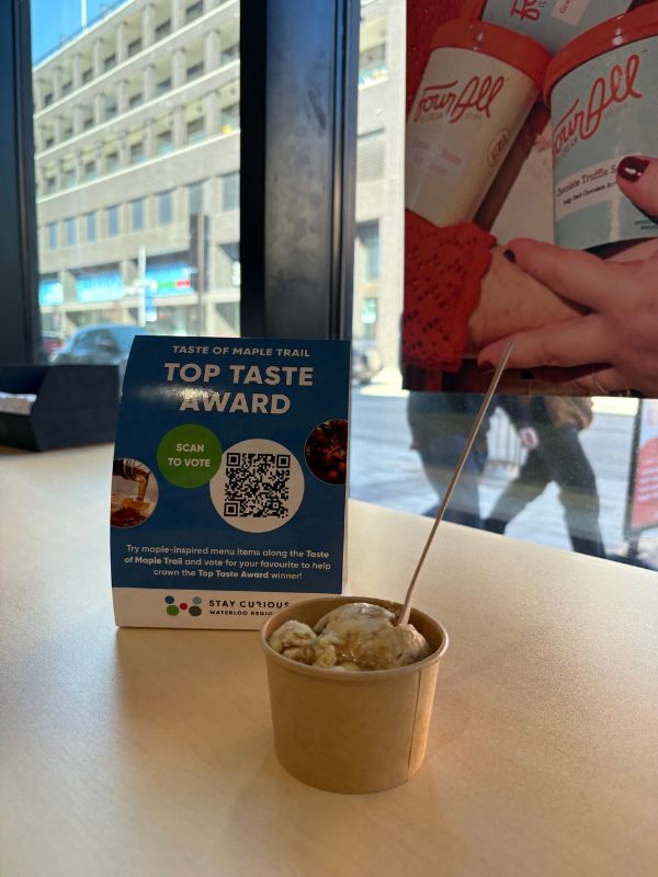 Inside the Kitchener location of Four All Ice Cream. There is a bowl of their Maple Brulee ice cream on a table beside a tent card promoting the Taste of Maple Trail Top Taste award encouraging people to vote for their favourite flavour. 