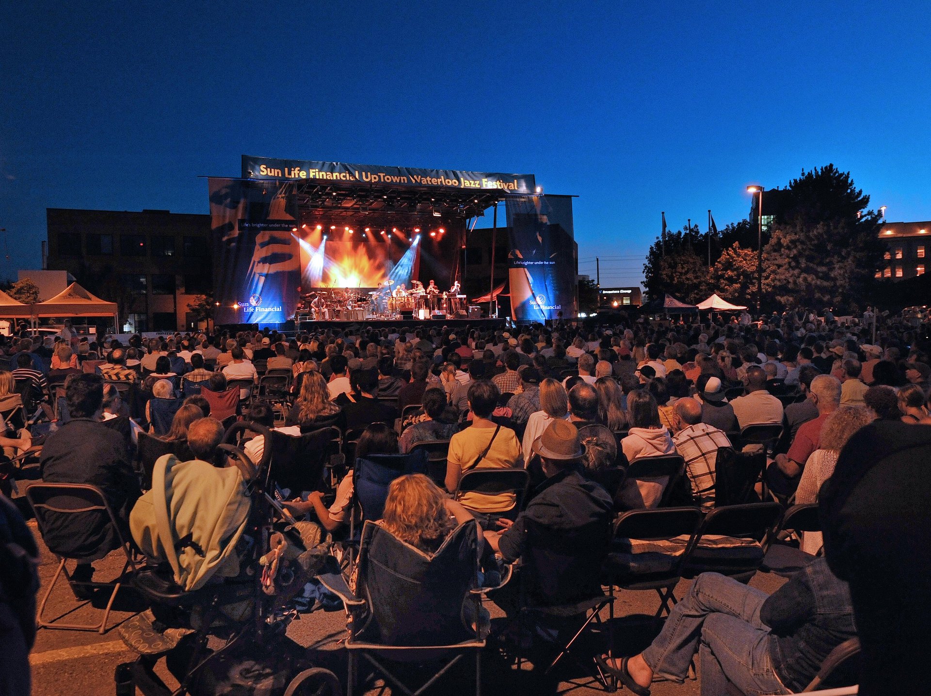An evening photo of the outdoor stage in Uptown Waterloo of the Uptown Waterloo Jazz Festival. There is a group performing on the stage, and a large crowd sitting in front of the stage taking in the performance.