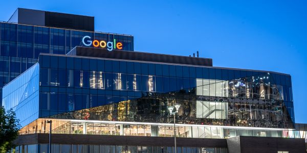 The outside of the Google building in the City of Waterloo. It is evening, and the Google word logo is lit up in its Google colours of blue, red, yellow, blue, green, red.