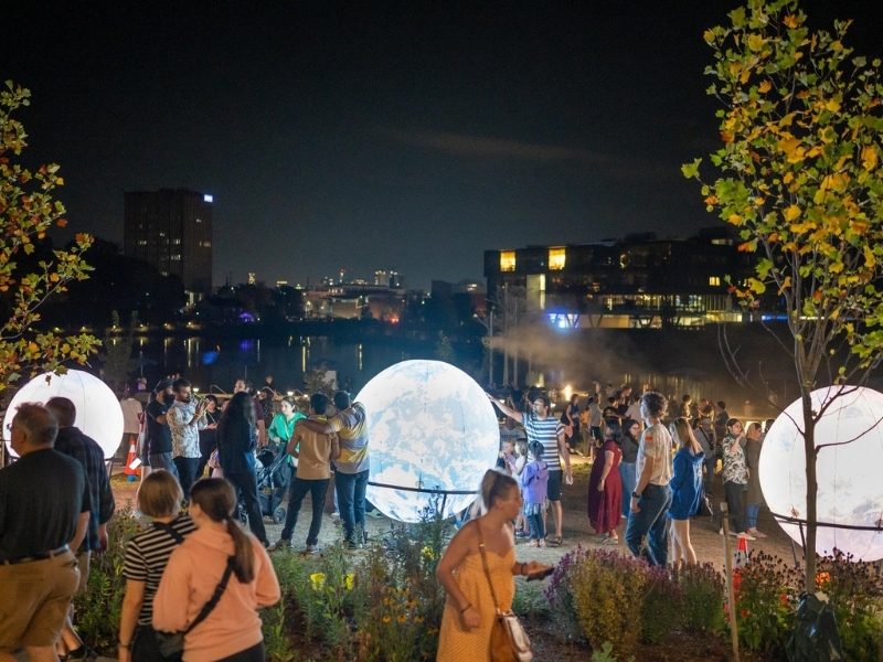 A large crowd of people taking in one of the Lumen Festival displays on the boardwalk along Silver Lake in Waterloo. The display features brightly lit up planet-looking installations: it is night and the installations are glowing.