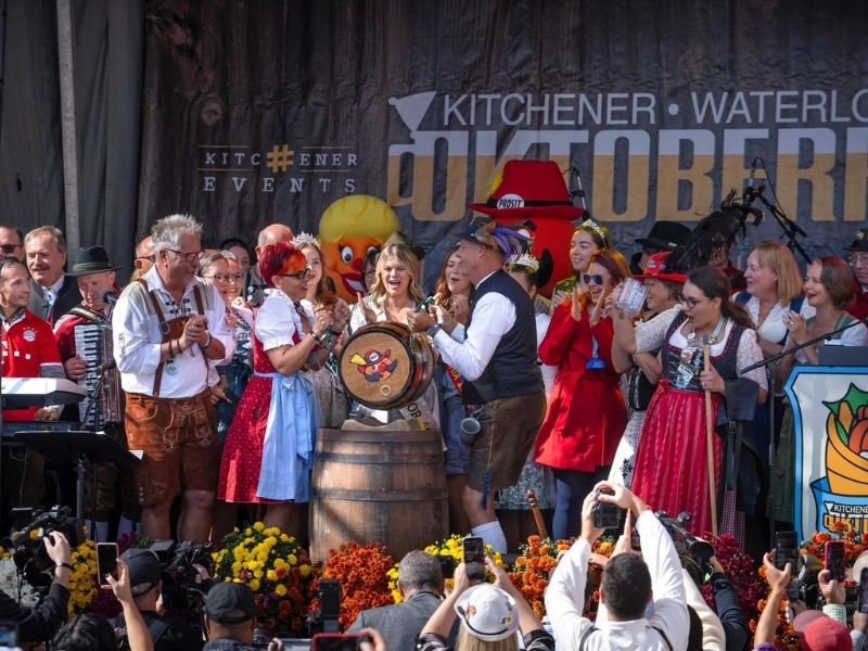 The main outdoor stage outdoors at Kitchener City Hall during the keg tapping for Kitchener-Waterloo Oktoberfest. There is a large crowd of dignitaries and Oktoberfest committee members, all wearing traditional Bavarian tracht, on stage getting ready to tap the keg.