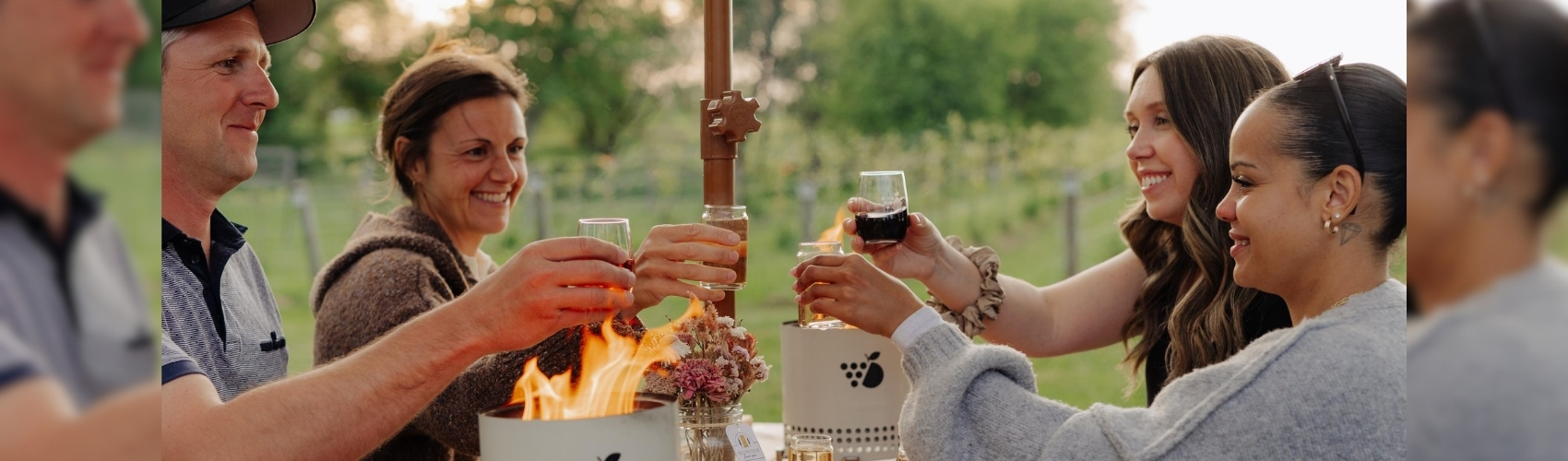 Four people sitting at a picnic table outdoors at The Perth Farmhouse. Tabletop fire bowls are lit on the table, and there are charcuterie boards set up. The group have beverages in hand and are doing a toast. It appears to be a fall evening.