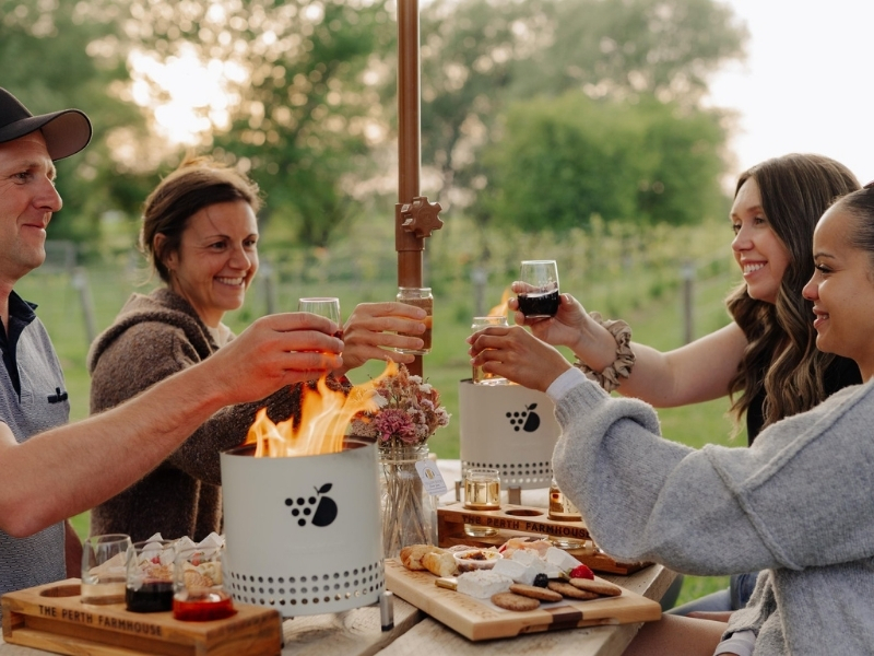 Four people sitting at a picnic table outdoors at The Perth Farmhouse. Tabletop fire bowls are lit on the table, and there are charcuterie boards set up. The group have beverages in hand and are doing a toast. It appears to be a fall evening.