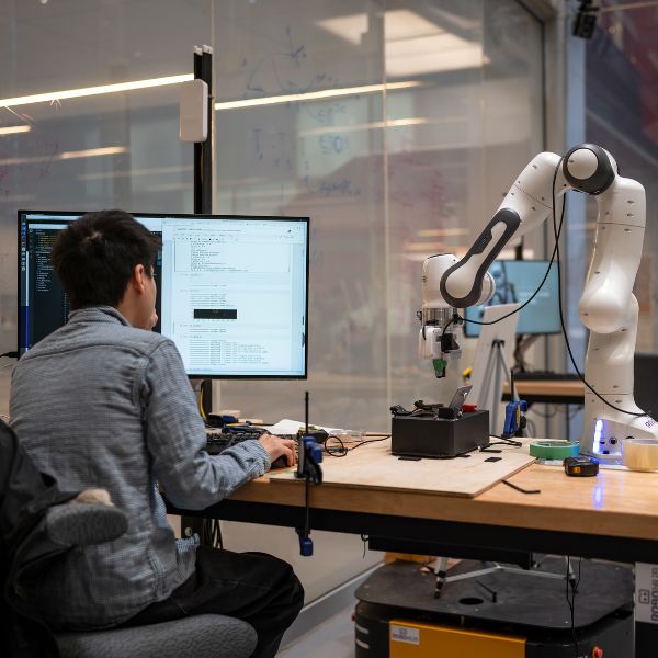 A person is seated at a desk in front of a computer. There is a robotic arm to their right, and they appear to be working on a program for it.