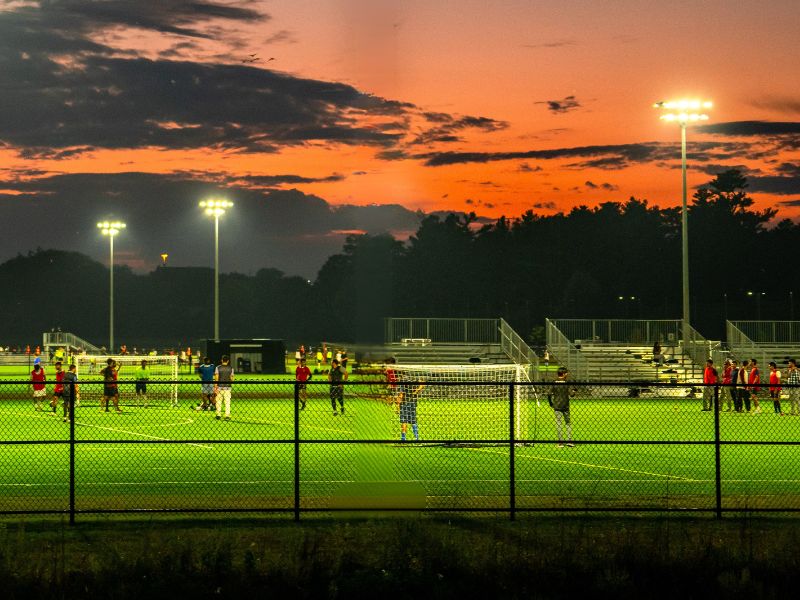A soccer field at twilight, with the sun setting in the background. You can see players on the field: the lights around the stadium are on and glowing.