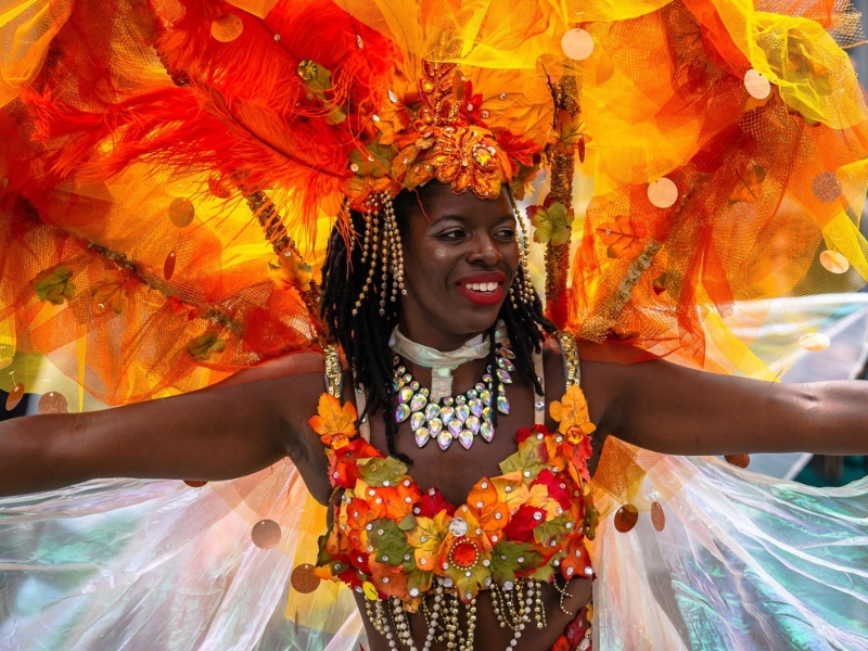 A Caribana Ignite participant in a colourful, orange feathered costume smiling at the crowd lining the parade route.