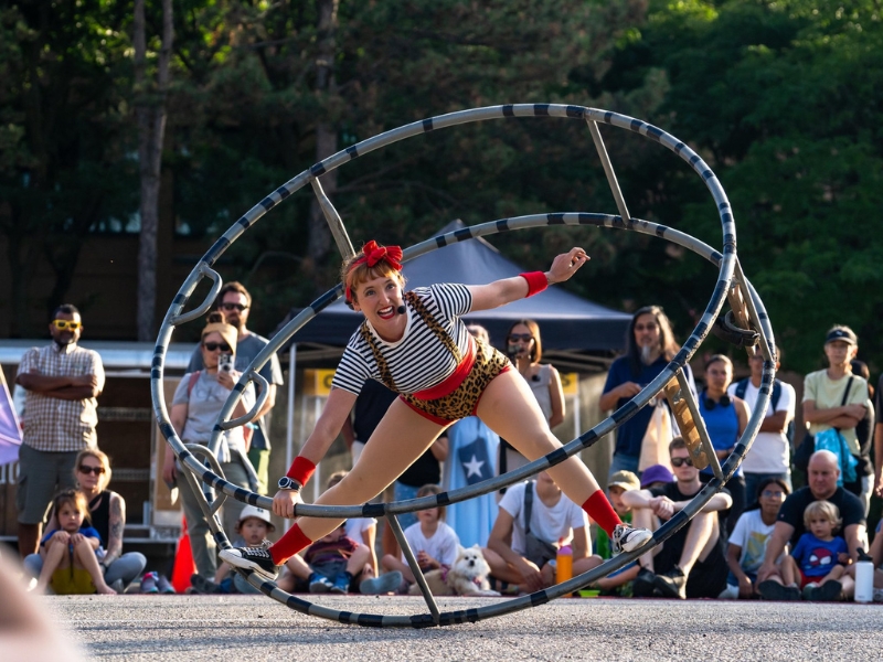 A busker is performing in a large circular metal wheel. A large crowd of spectators is watching the performance on the street in uptown Waterloo.