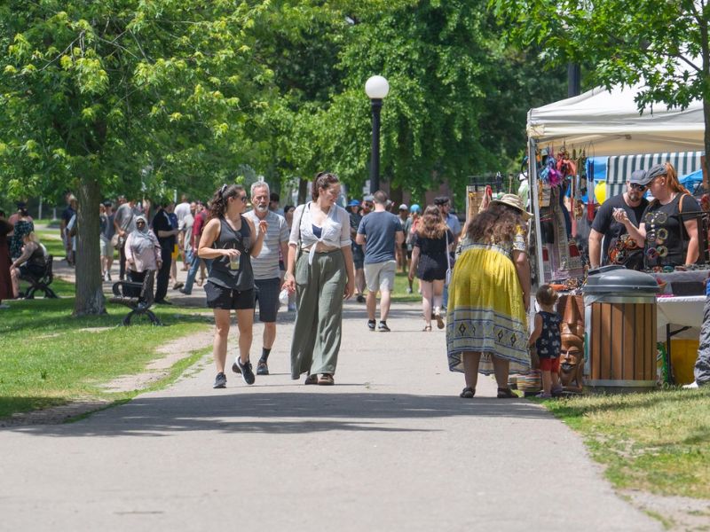 A crowd of people walking through Vicotria Park in Kitchener during the Multicultural Festival. It is a sunny summer's day, and you can see tented vendor stalls throughout the park.