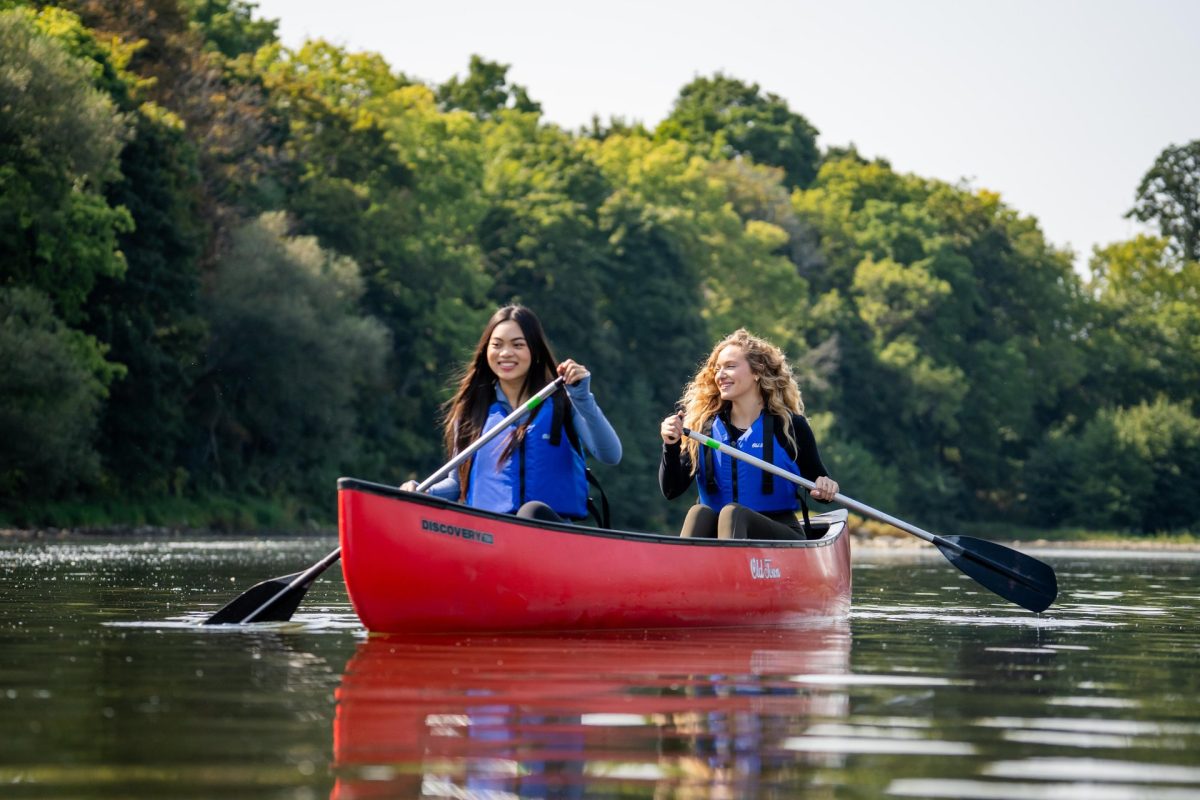 Two people in a canoe, paddling on the Grand River during the summer. It is a sunny, calm day on the water.