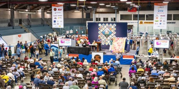 A large crowd inside the New Hamburg arena for the annual Quilt Auction that is part of the New Hamburg Mennonite Relief Sale. There are two large quilts being displayed on a raised stage by two traditionally-dressed Mennonite women, and an auctioneer taking bids.