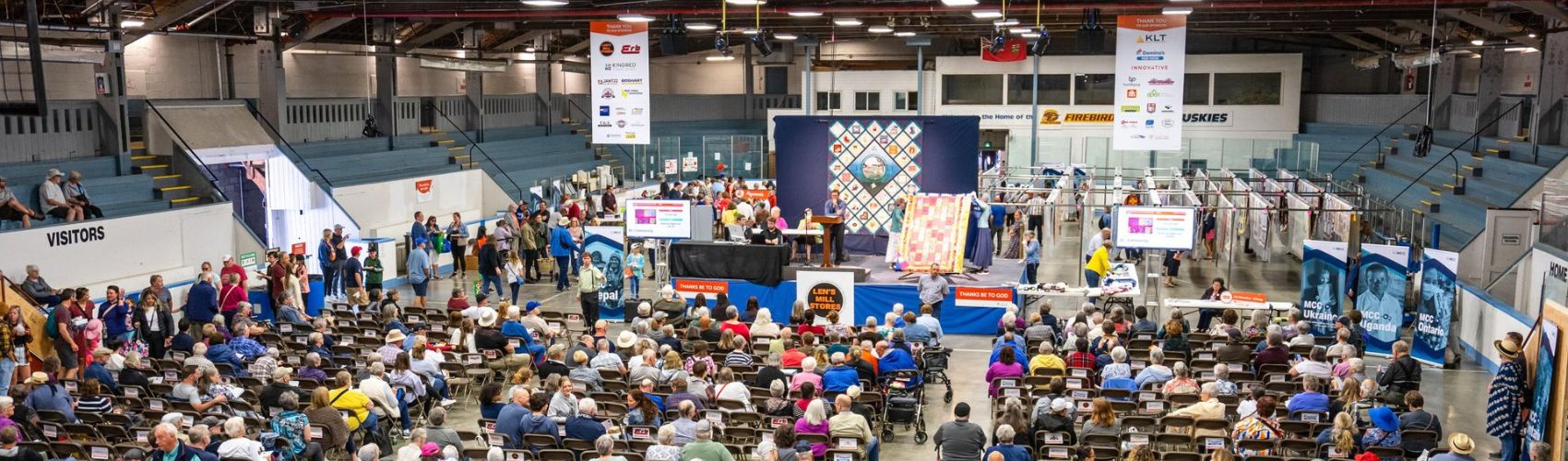 A large crowd inside the New Hamburg arena for the annual Quilt Auction that is part of the New Hamburg Mennonite Relief Sale. There are two large quilts being displayed on a raised stage by two traditionally-dressed Mennonite women, and an auctioneer taking bids.
