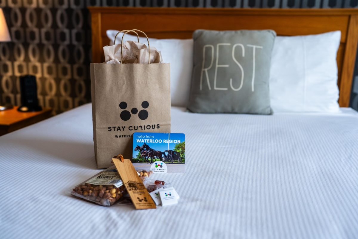 A bed in a room at the Walper Hotel in downtown Kitchener. There is a new Welcome Experience bag on the bed, with some of the contents arranged beside it. There is a decorative pillow with the word Rest on it also on the bed.