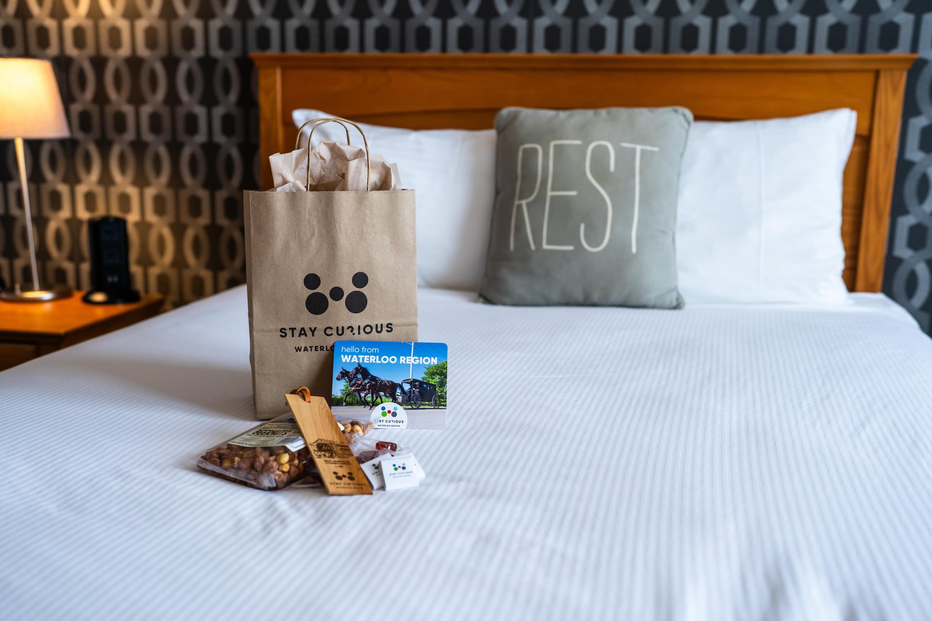A bed in a room at the Walper Hotel in downtown Kitchener. There is a new Welcome Experience bag on the bed, with some of the contents arranged beside it. There is a decorative pillow with the word Rest on it also on the bed. 