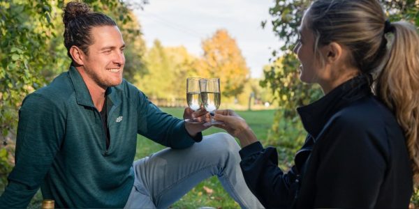 A couple sitting on the ground outdoors in a space that is surrounded by trees and green vegetation. They are enjoying a glass of wine, The sun is shining and they are smiling.