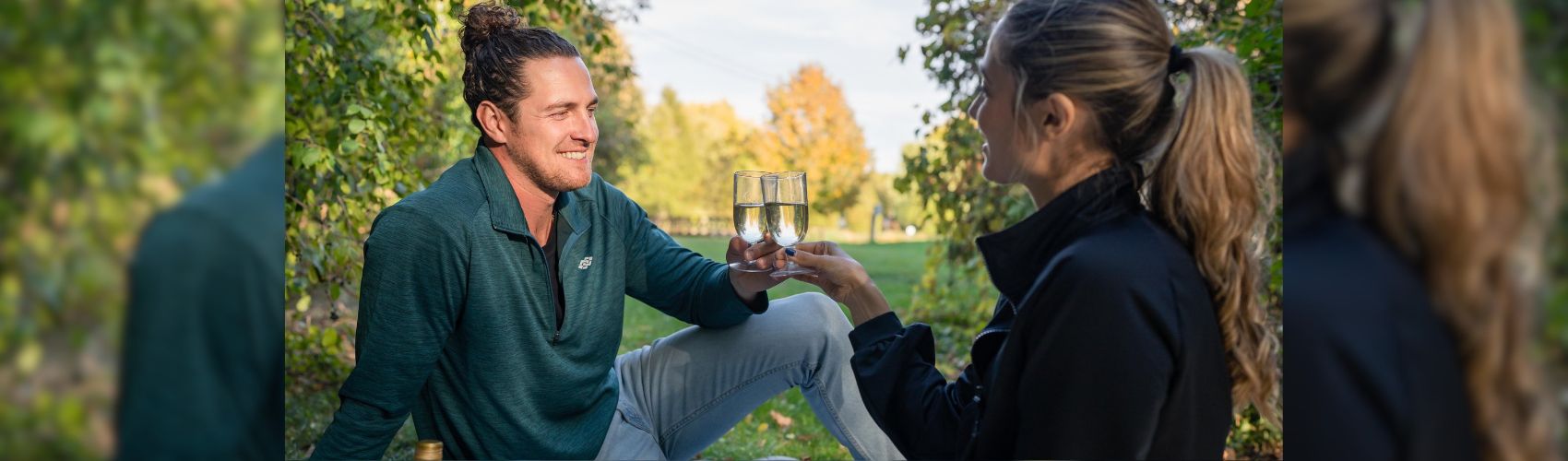 A couple sitting on the ground outdoors in a space that is surrounded by trees and green vegetation. They are enjoying a glass of wine, The sun is shining and they are smiling.