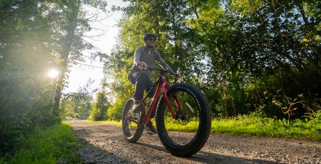 A person riding a fat bike on a gravel trail through a wooded area in Waterloo Region. It is summer and the tress are all green. The sun is shining through the trees.