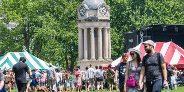 A crowd of people walking through Victoria Park in Kitchener during the annual Multicultural Festival. There are many vendor tents, and an outdoor stage at the base of the Clock Tower in the park.