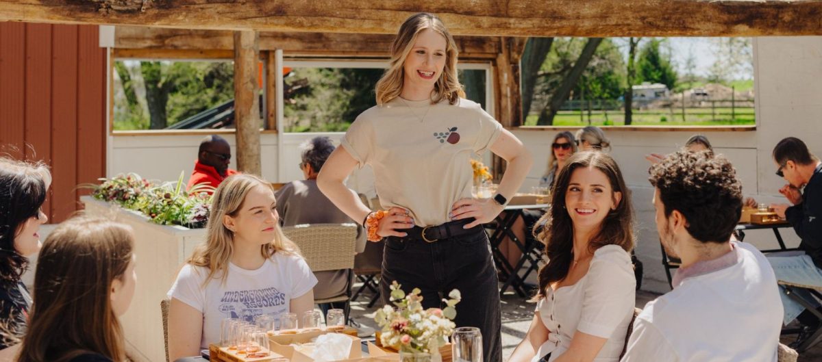 A staff member at The Perth Farmhouse is standing at an outdoor table where a group of people is seated. She is smiling, and appears to be speaking with the group. There are other tables in the pergola deck area, and all are enjoying a drink and something to eat.