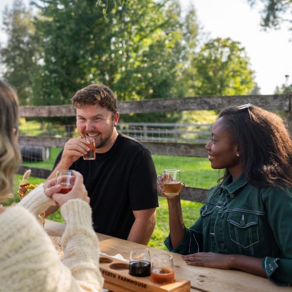 Three people are sitting around a picnic table outdoors at The Perth Farmhouse on a sunny summer's day. You can see fences and large green trees in the background. All three people are sampling drinks from a flight of beverages.