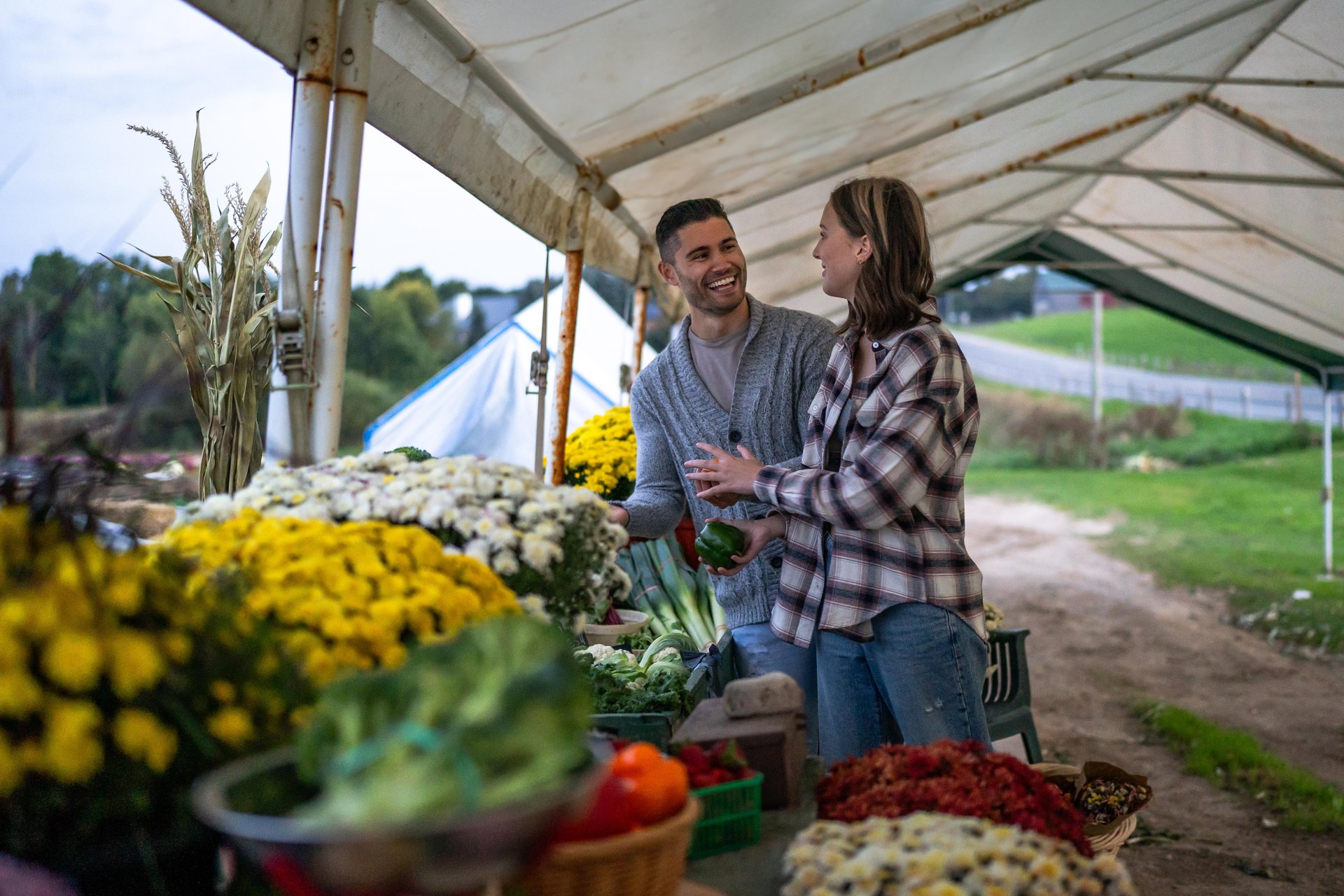 A couple going through various flowers and produce at a countryside farm gate stand. It appears to be a sunny fall day.