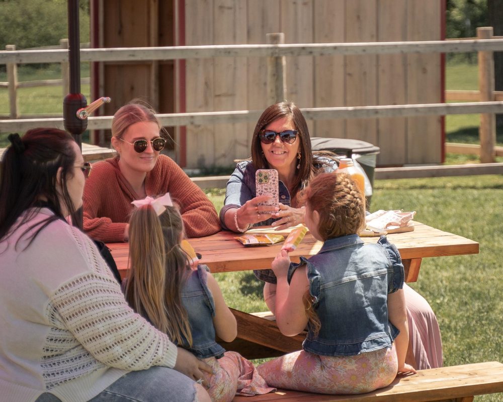 Three adults and two children sitting at a picnic table on a farm. One of the adults is smiling and holding a phone like she's taking a picture of the kids. It appears to be a sunny fall day.