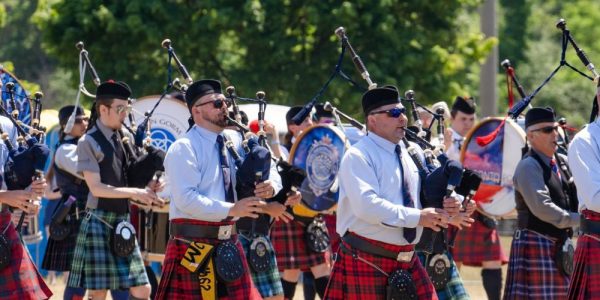 A parade of bag pipers all dressed in traditional Scottish kilts and garb. They are playing the pipes and parading as part of the Cambridge Scottish Games. It is a sunny summer's day.