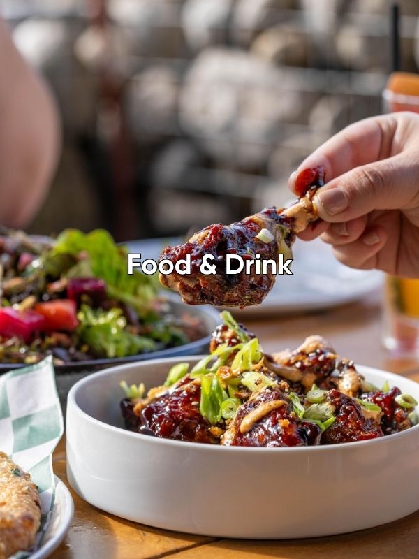 A white bowl filled with chicken drumsticks that appear to be covered in a teriyaki sauce, chopped green onions and toasted sesame seeds. A hand is holding one of the drumsticks above the bowl. The words overlayed on the image say Food and Drink