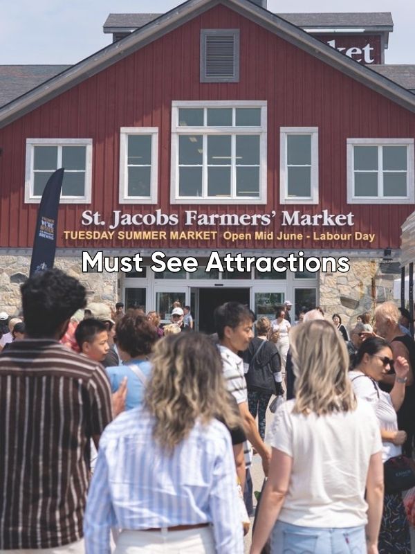 An exterior view of the main red building of the St. Jacobs Farmers' Market. It is summer, and there is a large crowd of people going into the market building. The words overlayed onto the image say Must Visit Attractions. 
