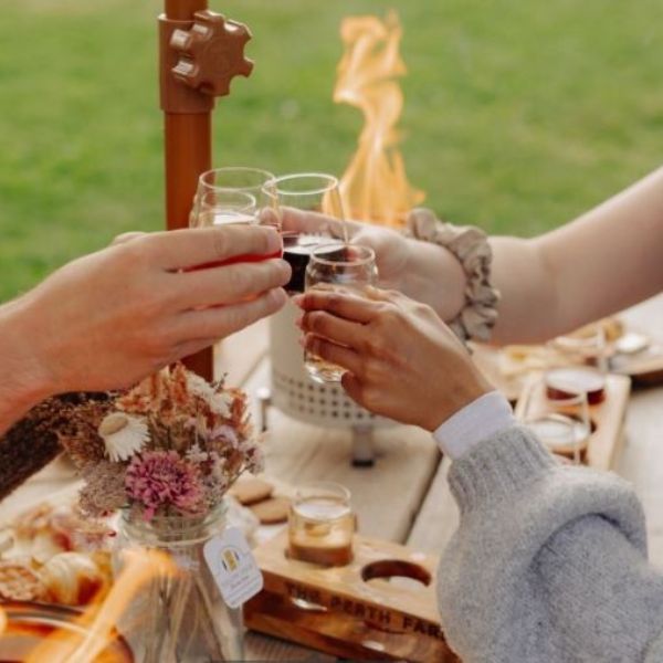 a close up of 3 hands holding beverage glasses that are clinking the cups together like a 'cheers'. There are wooden platters of food on the table. They appear to be outdoors at a picnic table in the summer: there is a small table top fire pot that is lit on the table. You can see the flames.