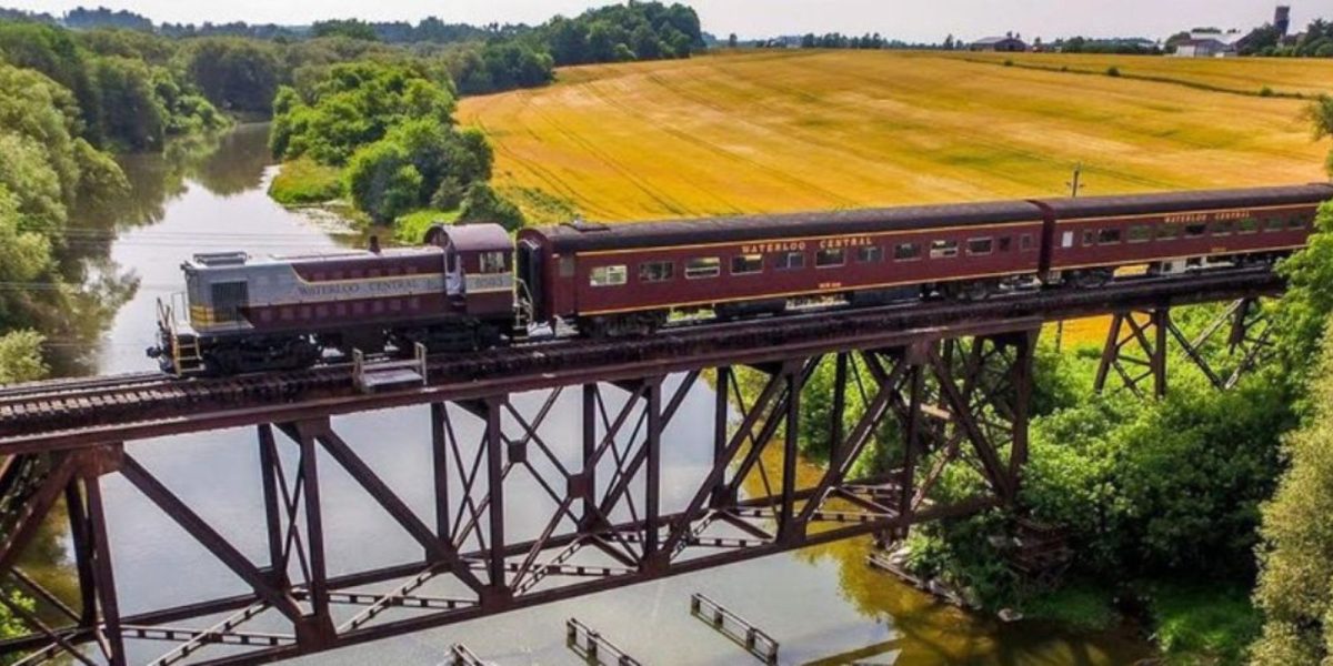 A Waterloo Central Railway train going across the tressle bridge over Conestogo River is St. Jacobs. You can see fields and trees on either side of the river.