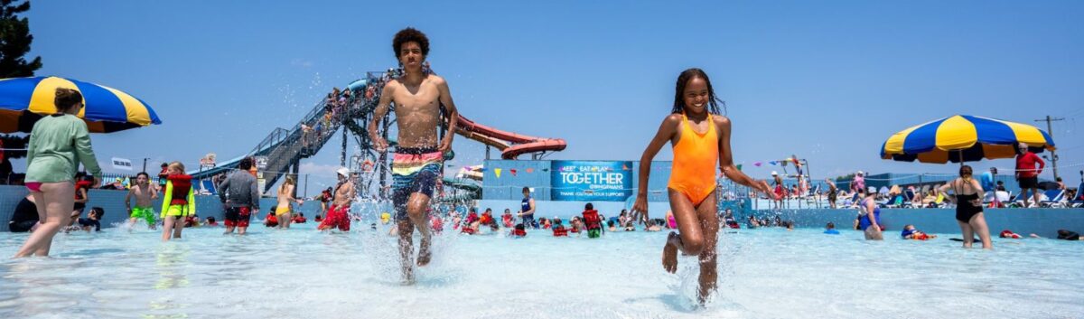 Two kids in bathing suits running towards the camera at the Big Splash Waterpark in Kitchener. There is a crowd of people enjoying the wave pool in the background. It is a sunny summer's day.