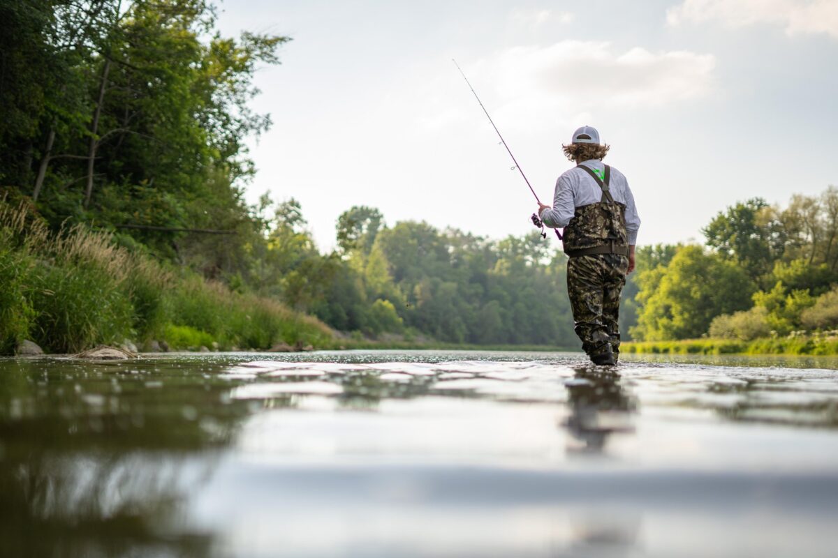 A person in hip waders fly fishing in the Grand River on a sunny summer's day. The banks of the river are lined with green trees and bushes.