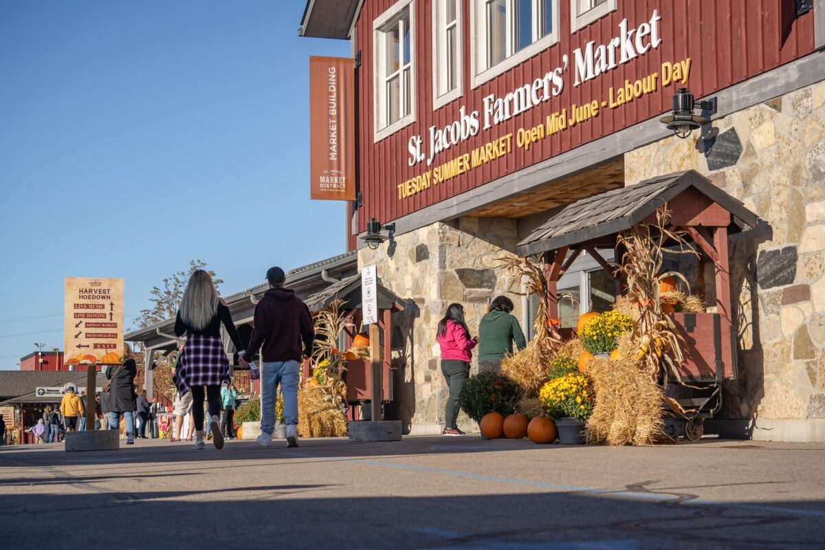 The exterior of the red main building of the St. JAcobs Farmers' Market. There is fall decor including straw bales, corn stalks and pumpkins decorating the main doors . People are walking into the building, as well as around the market grounds.