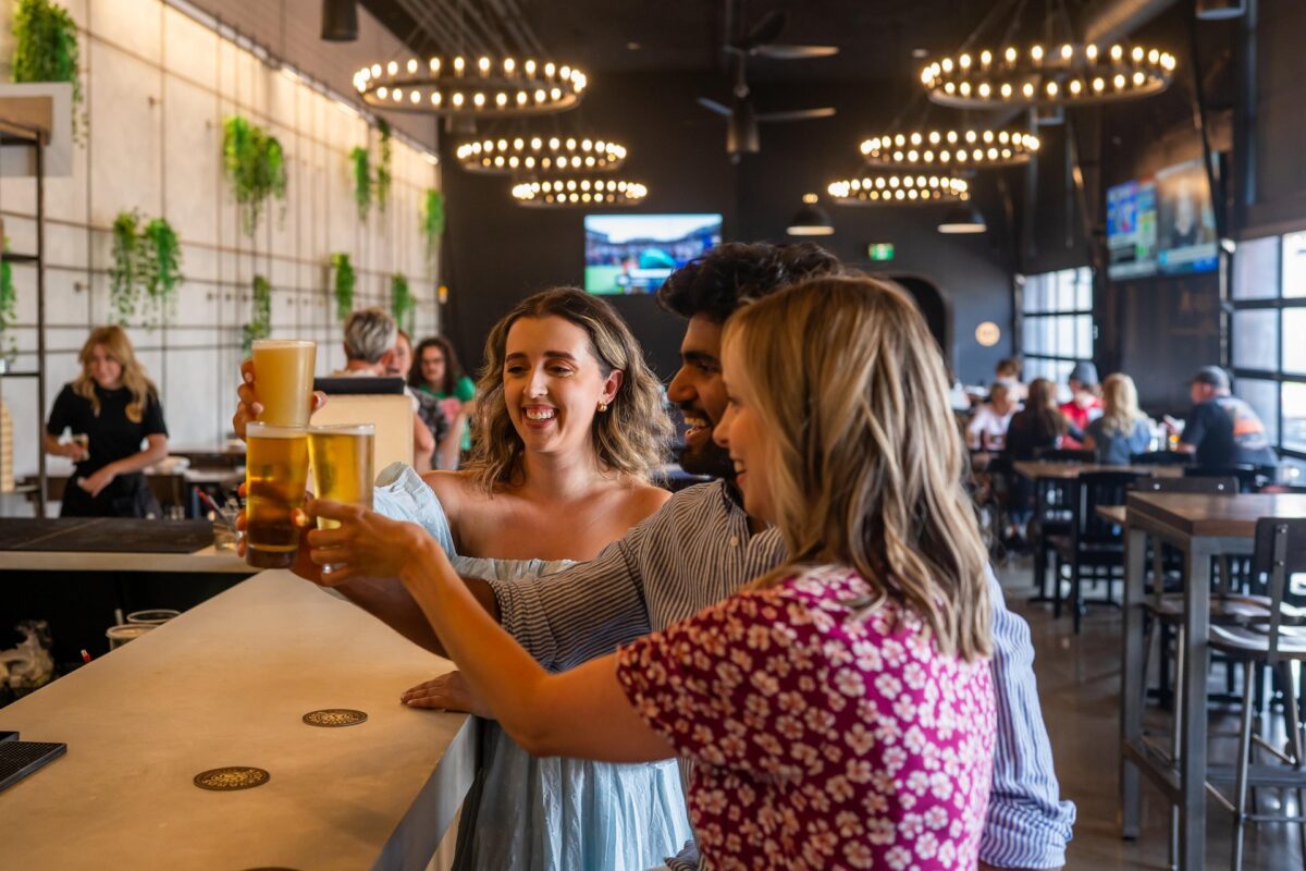 Three people lifting their glasses in a Cheers manner and smiling at the bar area of Stock Yards Brew Hall in the Market District of St. Jacobs.