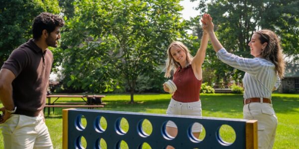 Three friends are playing a giant Connect Four game outdoors in the country on a sunny summer's day. Two of the friends are high-five-ing, like they have just made a great move.