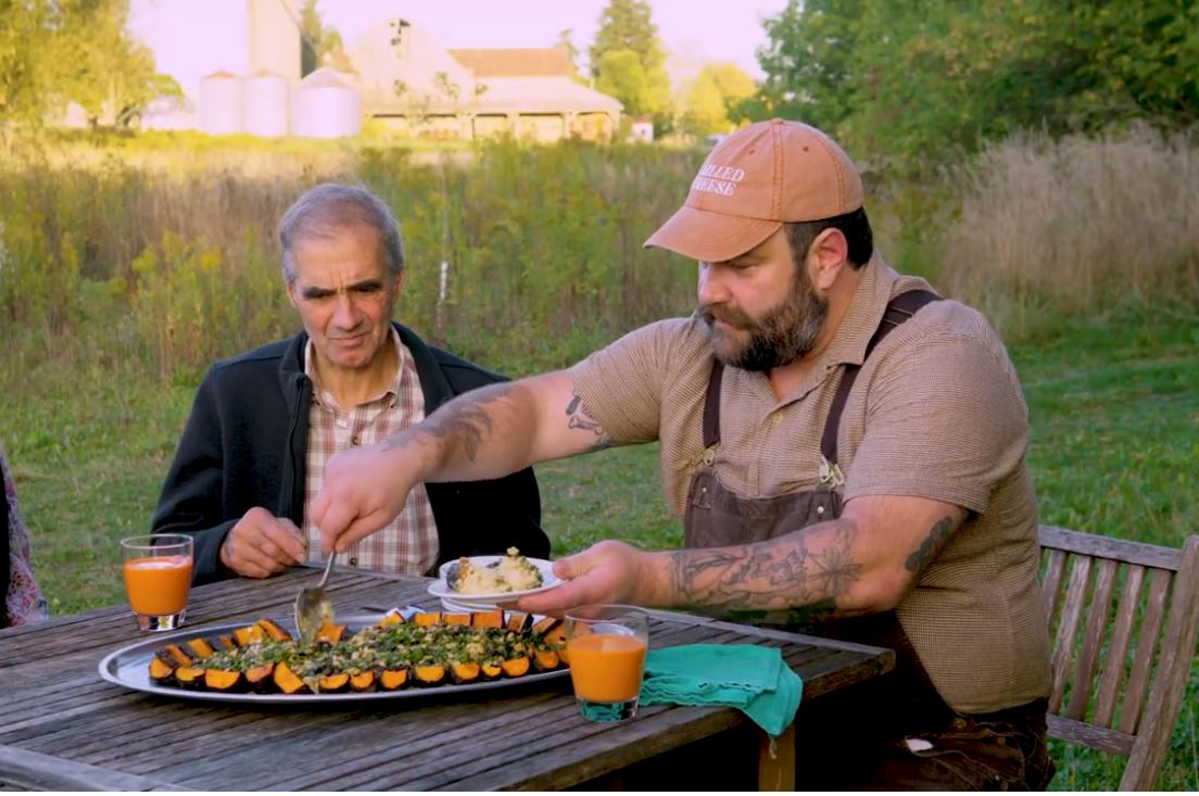 Host of Farm to Fork, Pfenings Organic, serving up a dish he created to a group who are seated outdoors at a farm.