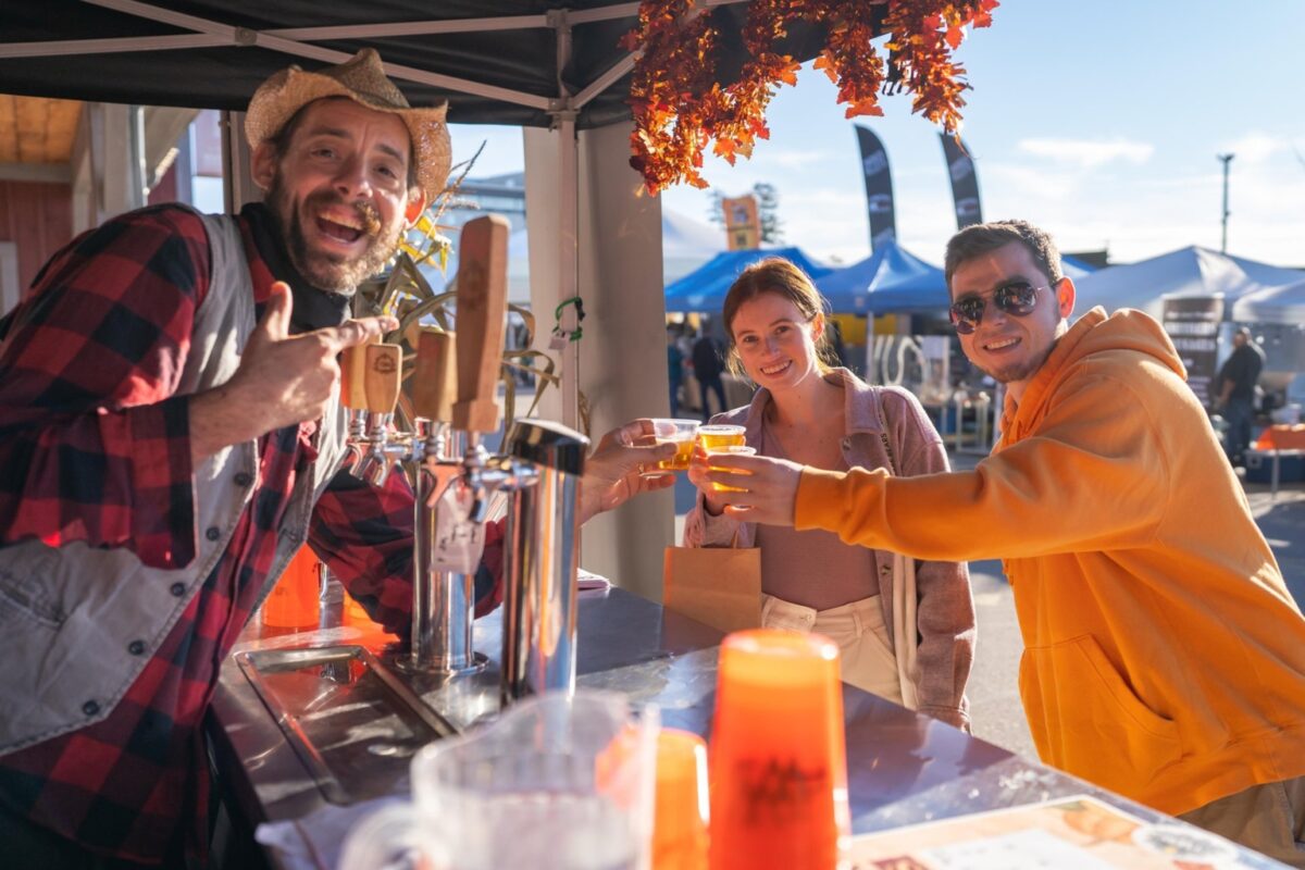 Three people doing cheers with glasses of craft beverages at one of the outdoor stalls at the St. Jacobs Farmers' Market. It is fall, and the booth is decorated with colourful leaves and other fall items. Everyone is smiling.