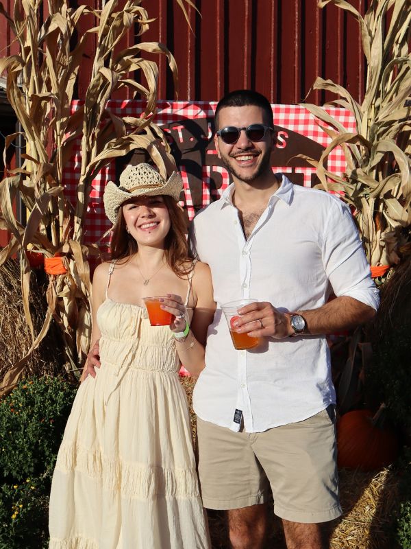A couple posing in front of a fall display of straw bales, pumpkins and corn stalks on a sunny fall day. They are smililng and holding beverages.