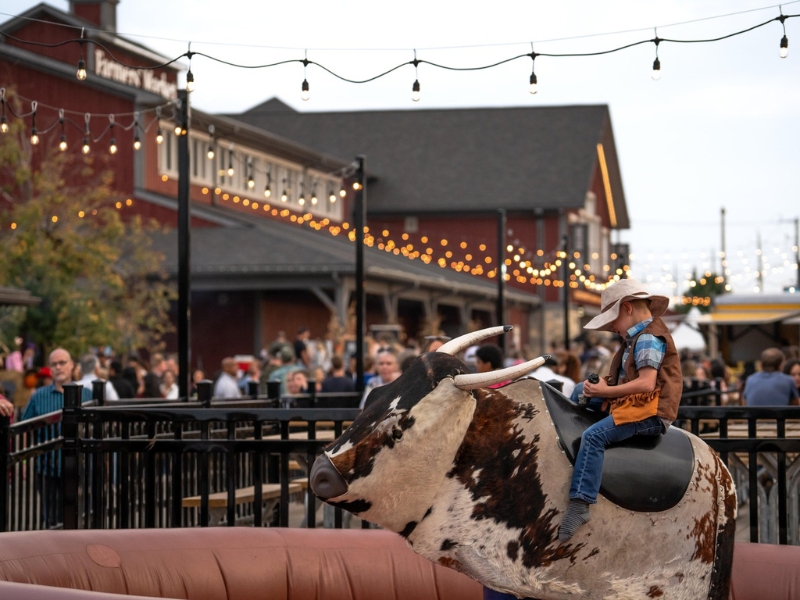 The main red building of the St. Jacobs Farmers' Market during their Harvest Hoedown event. There are edison lights strung up along an outdoor seating area. You can see a large crowd milling about, and a boy in a cowboy hat seated on a mechanical bull in the forefront.