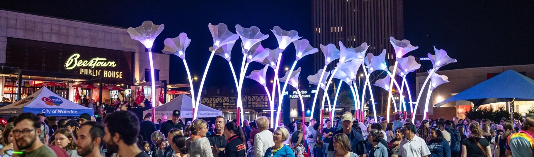 A large crowd of people walking through an illuminated display in Waterloo Square at night, that is part of the annual Lumen Festival in Uptown Waterloo.