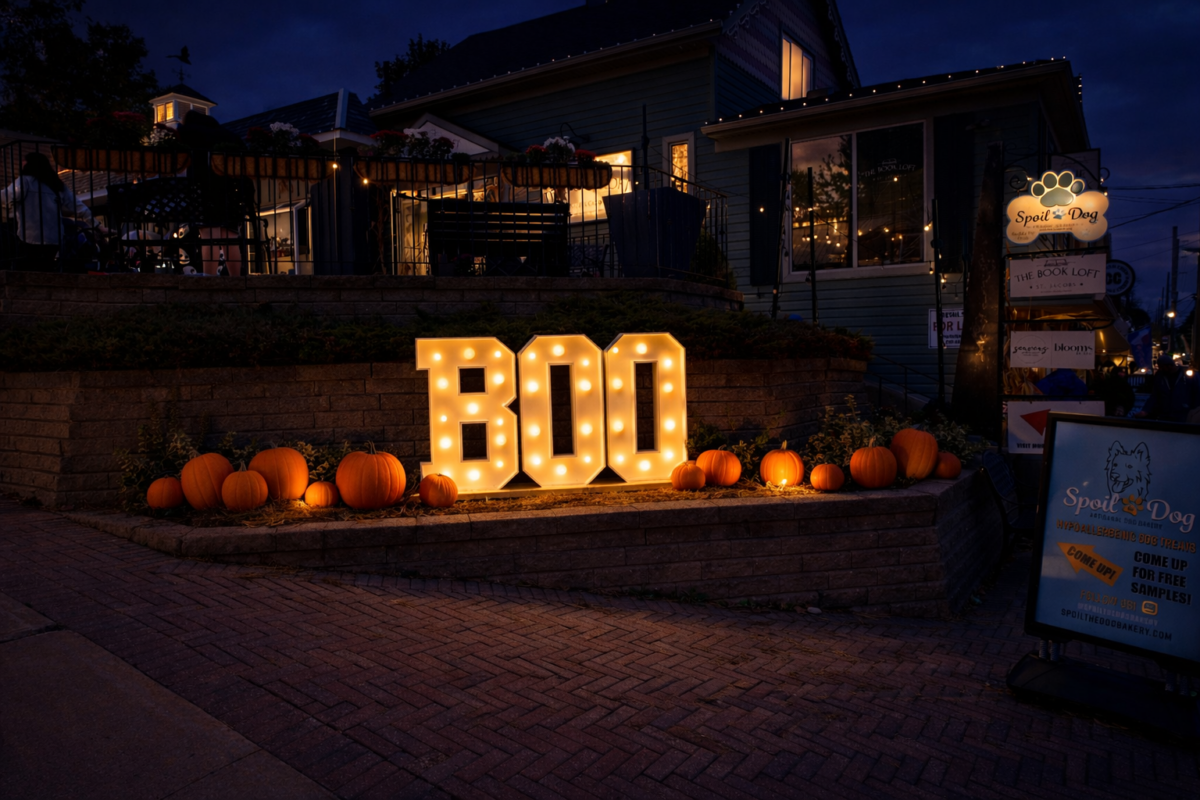 A night time shot in the Village of St. Jacobs during the annual Pumpkinpalooza event. There are any carved pumpkins sitting on straw bales in front of the shops, and a lit-up sign that spells the word BOO. 