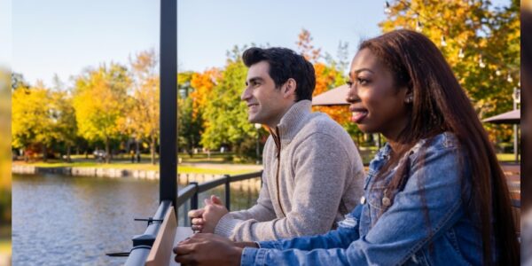 A couple leaning on a railing of a patio overlooking the water in Kitchener's Victoria Park. It is a sunny fall day: you can see the changing colours on the trees around the water.