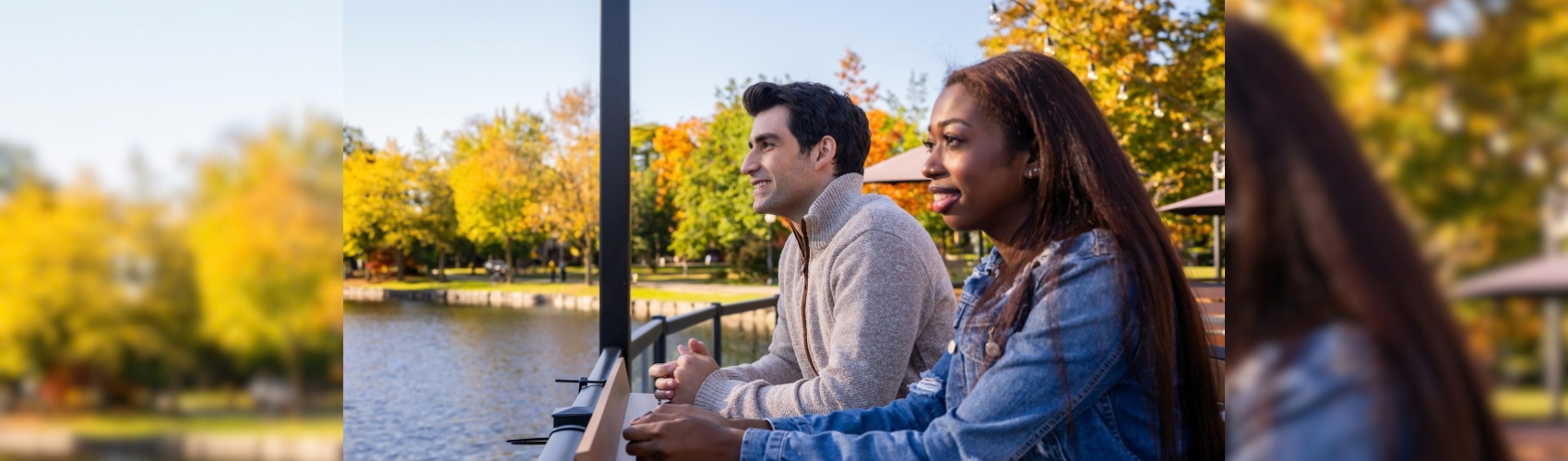 A couple leaning on a railing of a patio overlooking the water in Kitchener's Victoria Park. It is a sunny fall day: you can see the changing colours on the trees around the water.