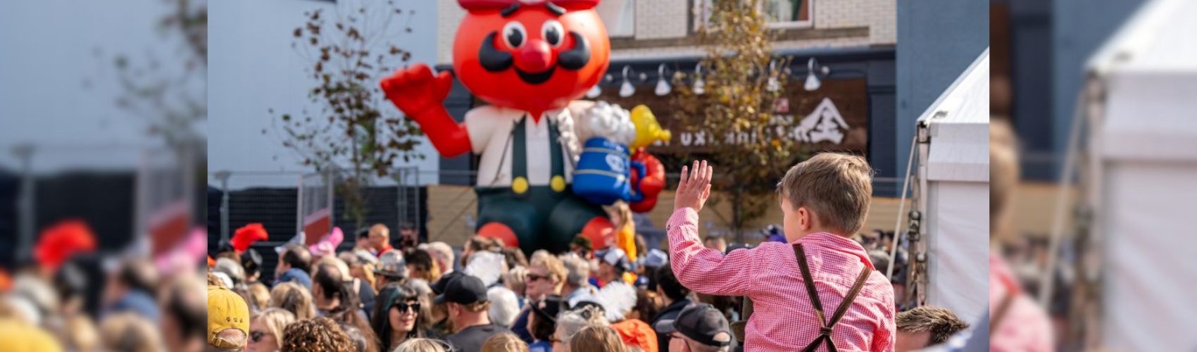 A young child in traditional Bavarian tracht is watching the keg tapping at the opening ceremony of Oktoberfest. The child is sitting on their parents' shoulders: there is a large crowd outdoors at the ceremony.